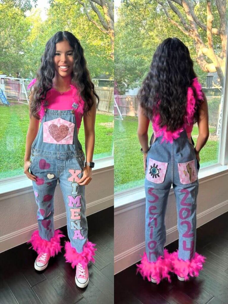 A young woman wearing decorated denim overalls for a senior year celebration. The overalls are embellished with pink fringe, hearts, and the name 'XIMENA' down one leg. The back legs display the words 'Senior' and '2024'.
