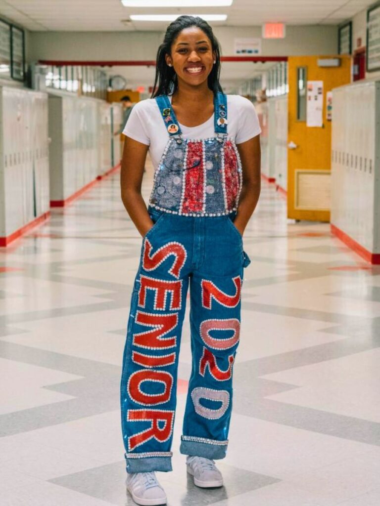 A smiling young woman stands in a school hallway wearing custom-decorated denim overalls. The overalls are embellished with 'SENIOR' and '2020' in bold red and white letters down the legs.