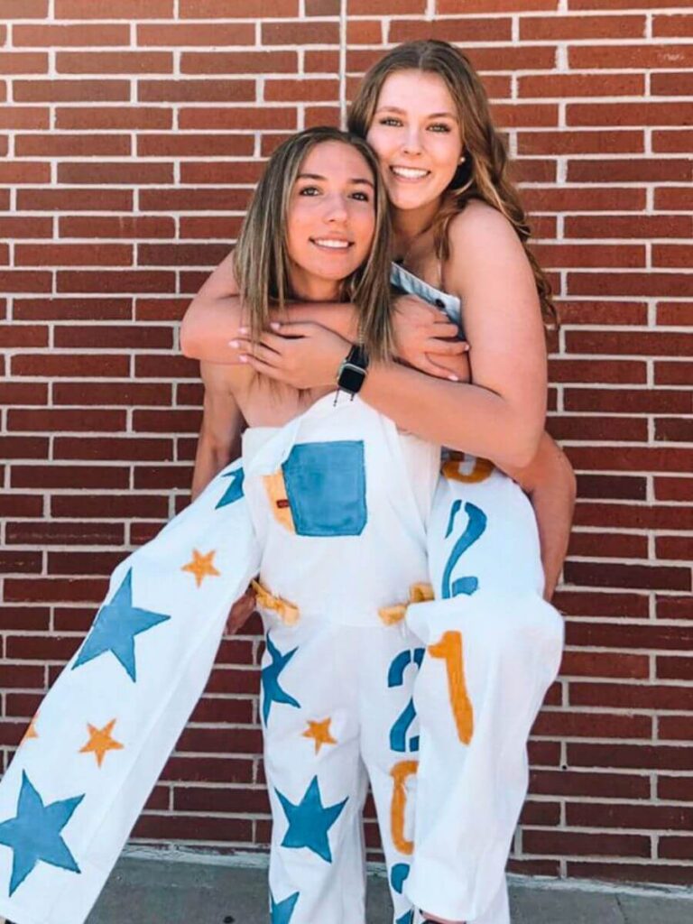 Two happy young women posing in front of a brick wall. Both wearing white overalls painted with blue and orange stars.