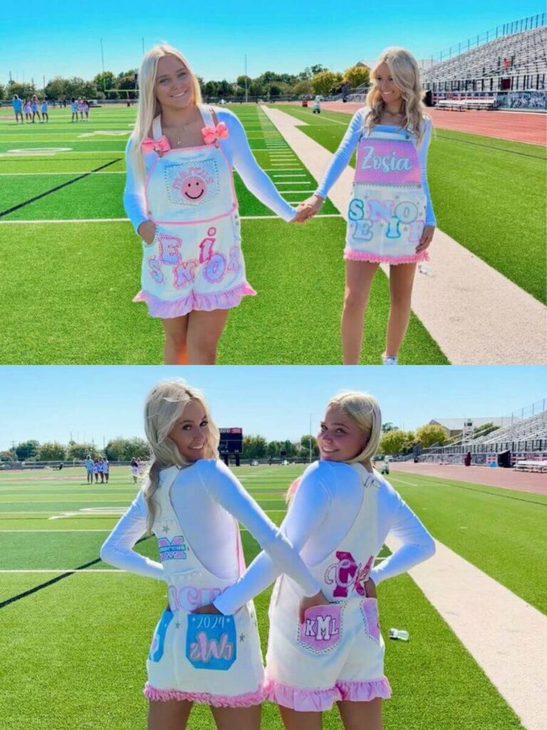 Two high school seniors stand hand-in-hand on a football field, wearing creatively customized white overalls with pink trim and colorful designs.