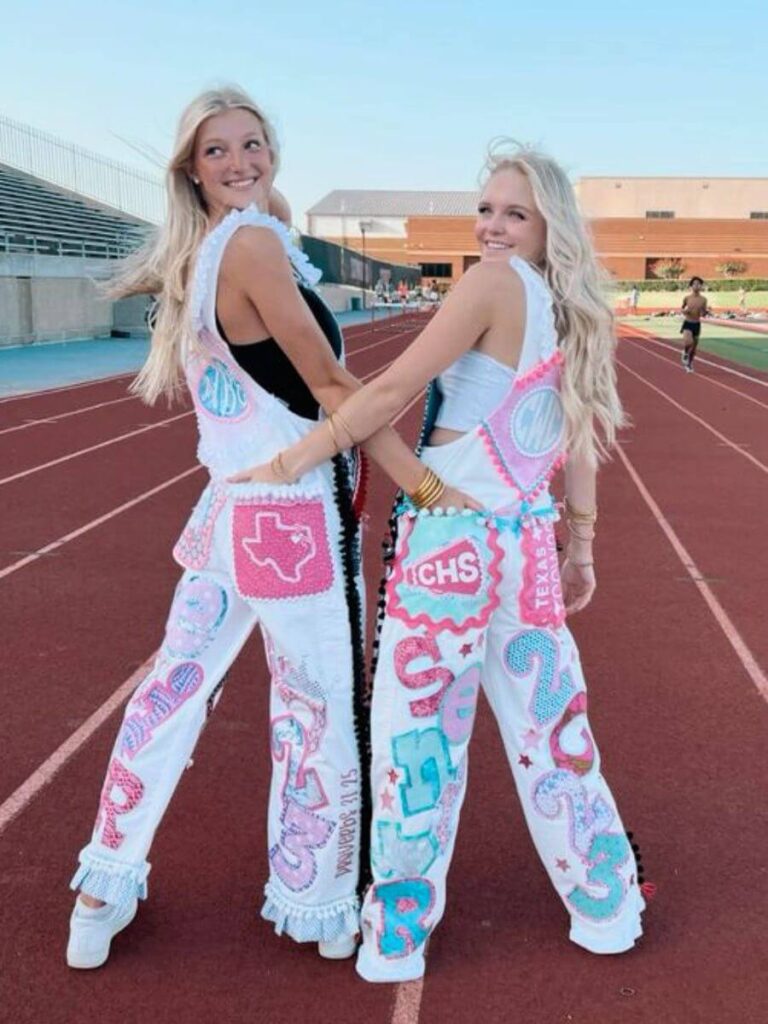 Two smiling high school seniors on a track field, linked arm in arm, displaying the back of their custom-decorated overalls. The overalls are adorned with various designs, including the word 'SENIOR', stars, hearts, and their school initials 'RCHS', all in a mix of pink, blue, and white.