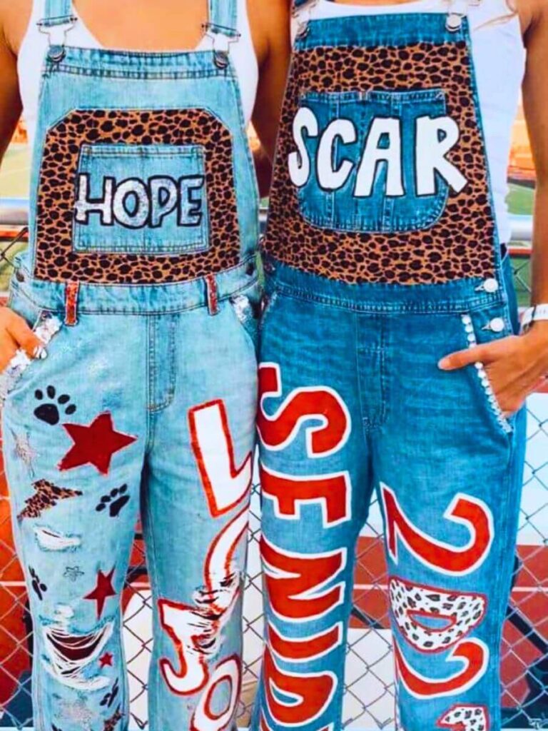 Two people wearing customized denim overalls with large letters spelling out 'HOPE' and 'SCAR' on the bibs, surrounded by leopard print patterns. The legs are painted with the words 'SENIOR' in bold red, along with paw prints and stars.