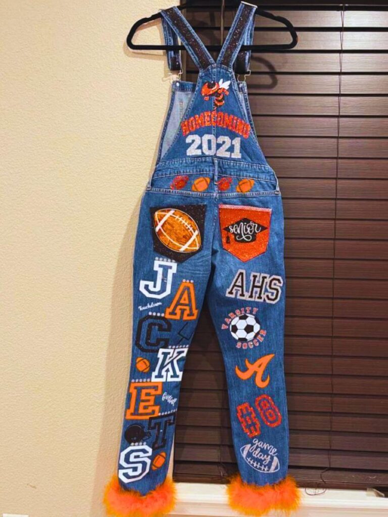 Custom-painted senior overalls hanging against a wooden background, with 'Homecoming 2021' written across the top. The legs are adorned with school spirit motifs including the word 'JACKETS', footballs, a volleyball, and a soccer ball, as well as a school logo for AHS.