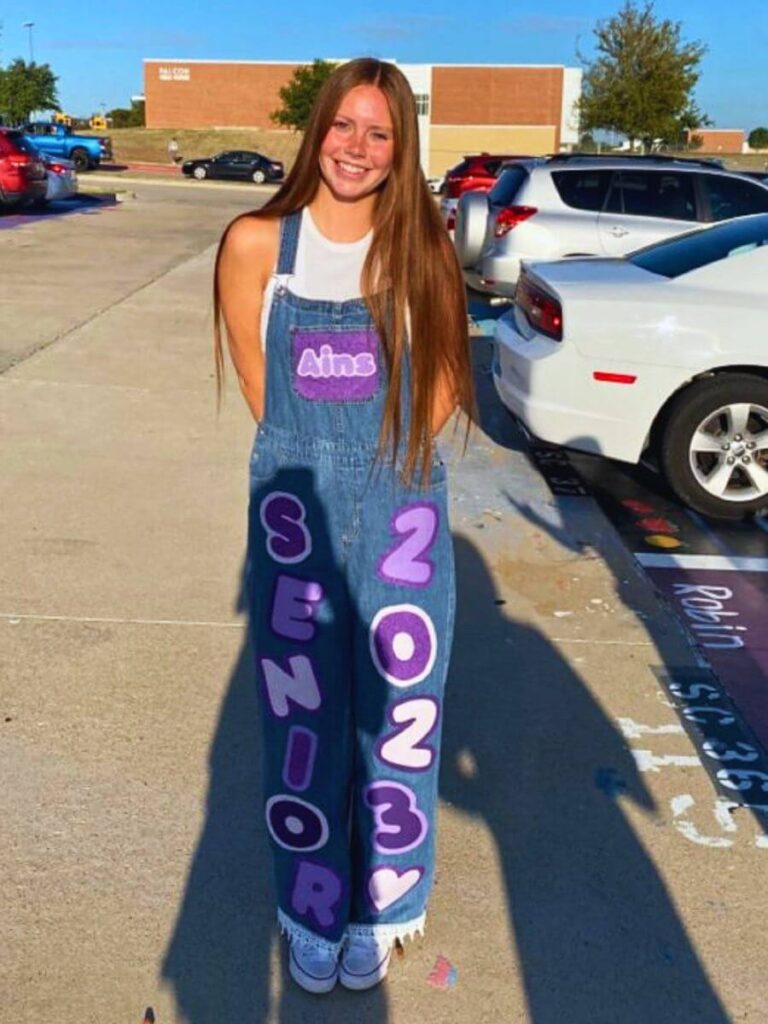 A smiling young woman stands in a parking lot, wearing customized denim overalls for senior year festivities. The overalls feature her name 'Aims' on the front bib in purple lettering and 'SENIOR' and '2023' down each leg.
