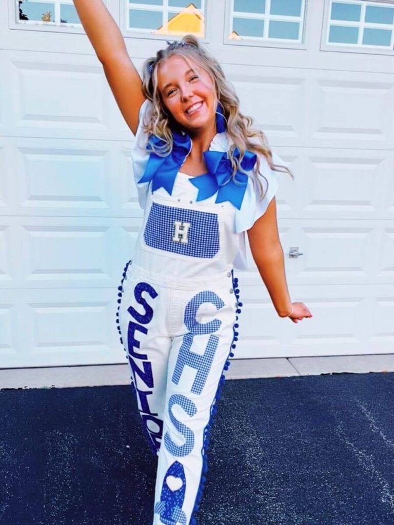 A young woman posing with one arm raised, wearing white overalls with 'SENIOR' and a school emblem in blue lettering down the leg. The overalls are embellished with blue ribbons.