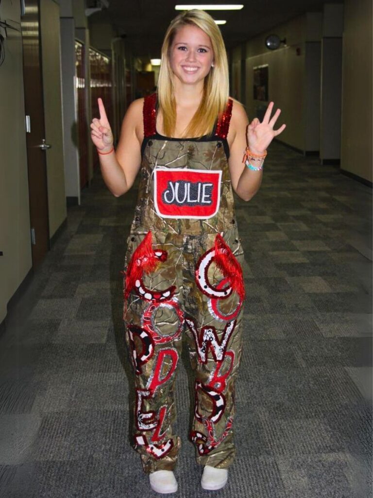 A cheerful young woman standing in a hallway, sporting customized camouflage overalls with her name 'JULIE' on the bib. The overalls are decorated with red and silver glittering accents and letters.