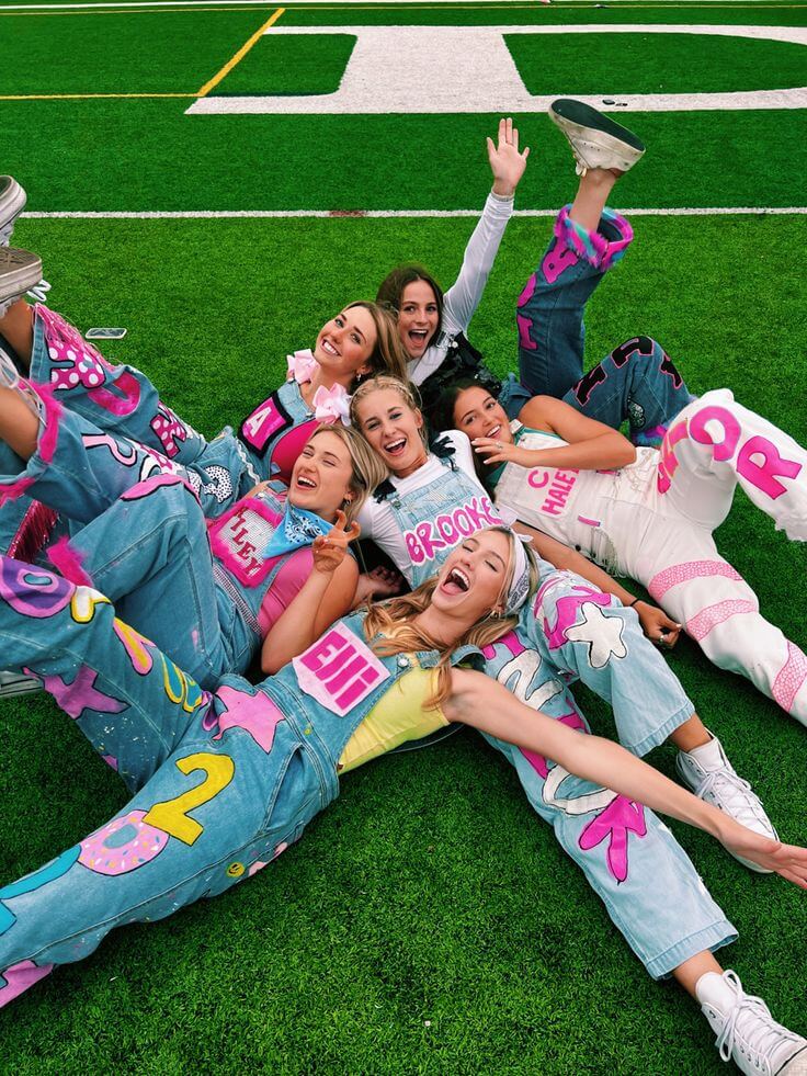 A joyful group of high school seniors lying on a football field, laughing and posing in their brightly decorated overalls.